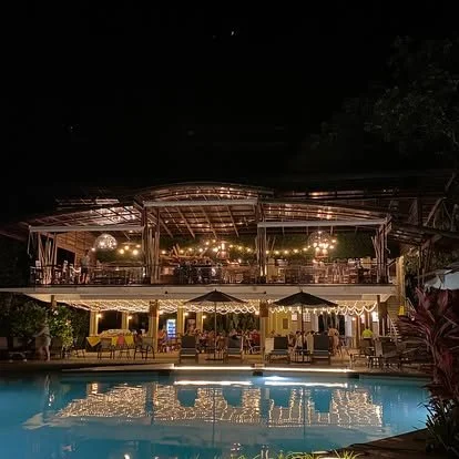 Night scene of an outdoor swimming pool with a bar and seating area, illuminated by string lights and lamps.