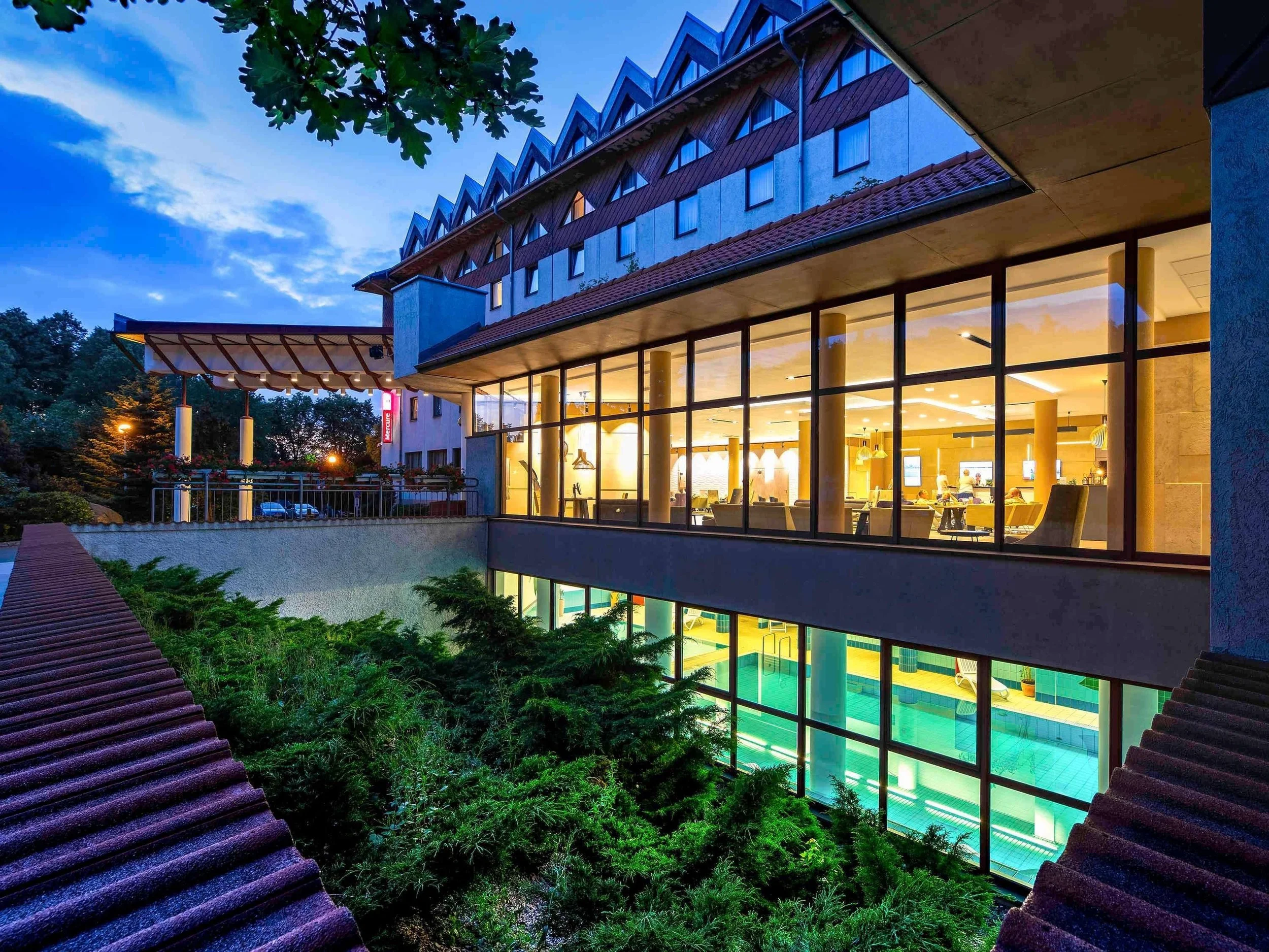 A multi-story hotel building with large glass windows showing a brightly lit indoor pool area, with outdoor greenery and a led sign on the corner of the building.