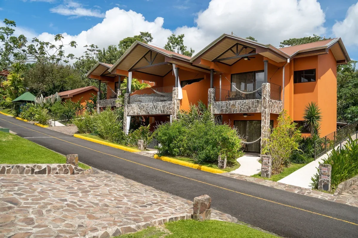 A two-story orange house with stone accents, surrounded by lush green plants, trees, and a driveway with a stone border, under a partly cloudy sky.