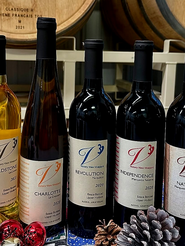 Four bottles of Victory View Vineyard wine, including Charlotte, Revolution, and Independence, displayed on a table with wine barrels in the background.