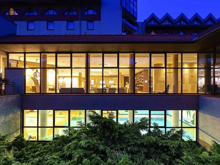 Nighttime view of a building with large glass windows revealing a lit interior, including a dining or meeting area on the upper floor and a brightly lit indoor pool on the lower floor, surrounded by greenery.