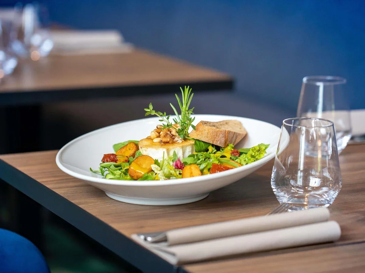 A plate of salad with mixed greens, roasted vegetables, cheese, bread slices, and garnished with herbs on a wooden table set for dining.