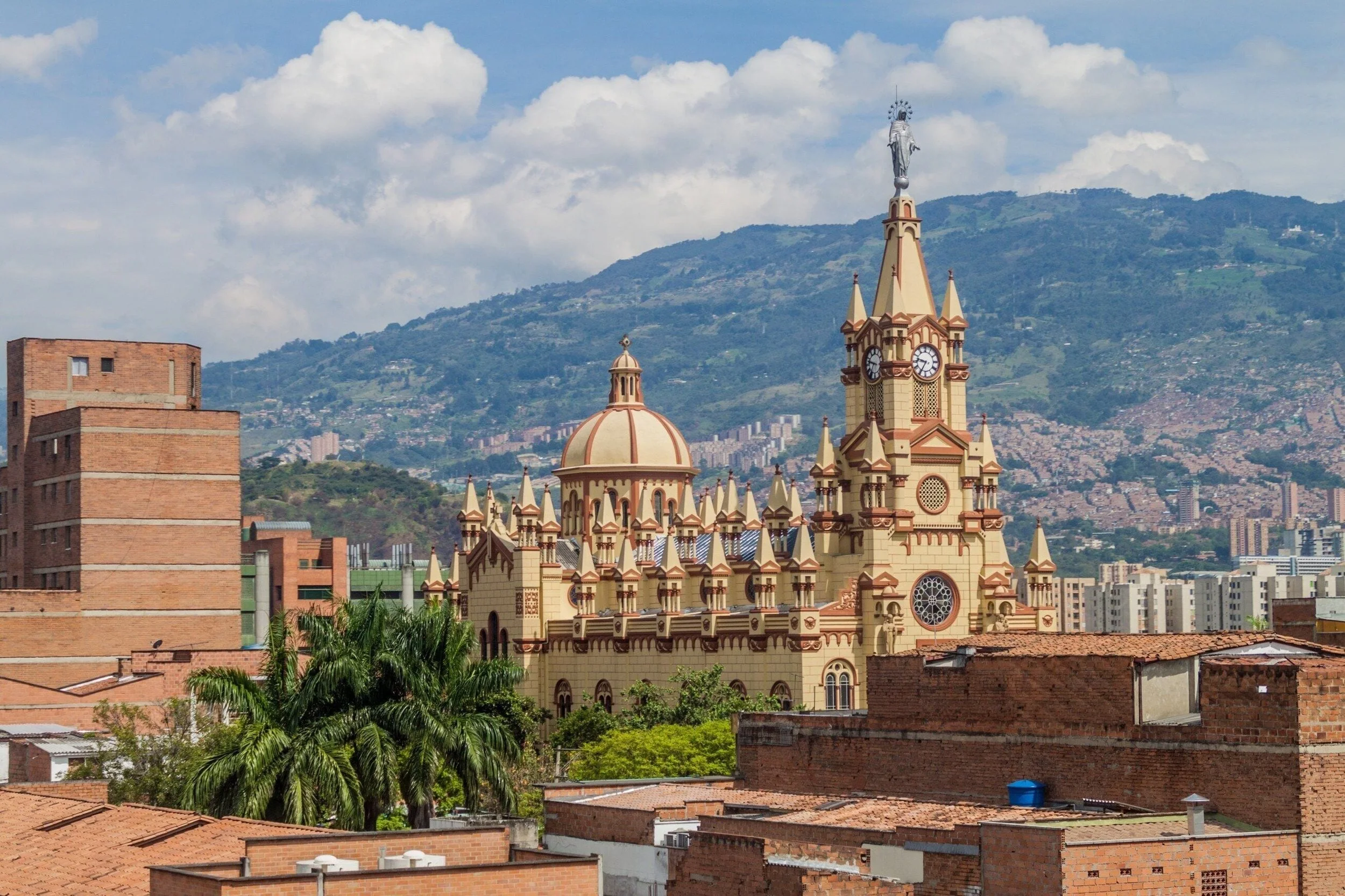 A cityscape with a historic church featuring a clock tower, situated against a backdrop of green mountains and a partly cloudy sky.