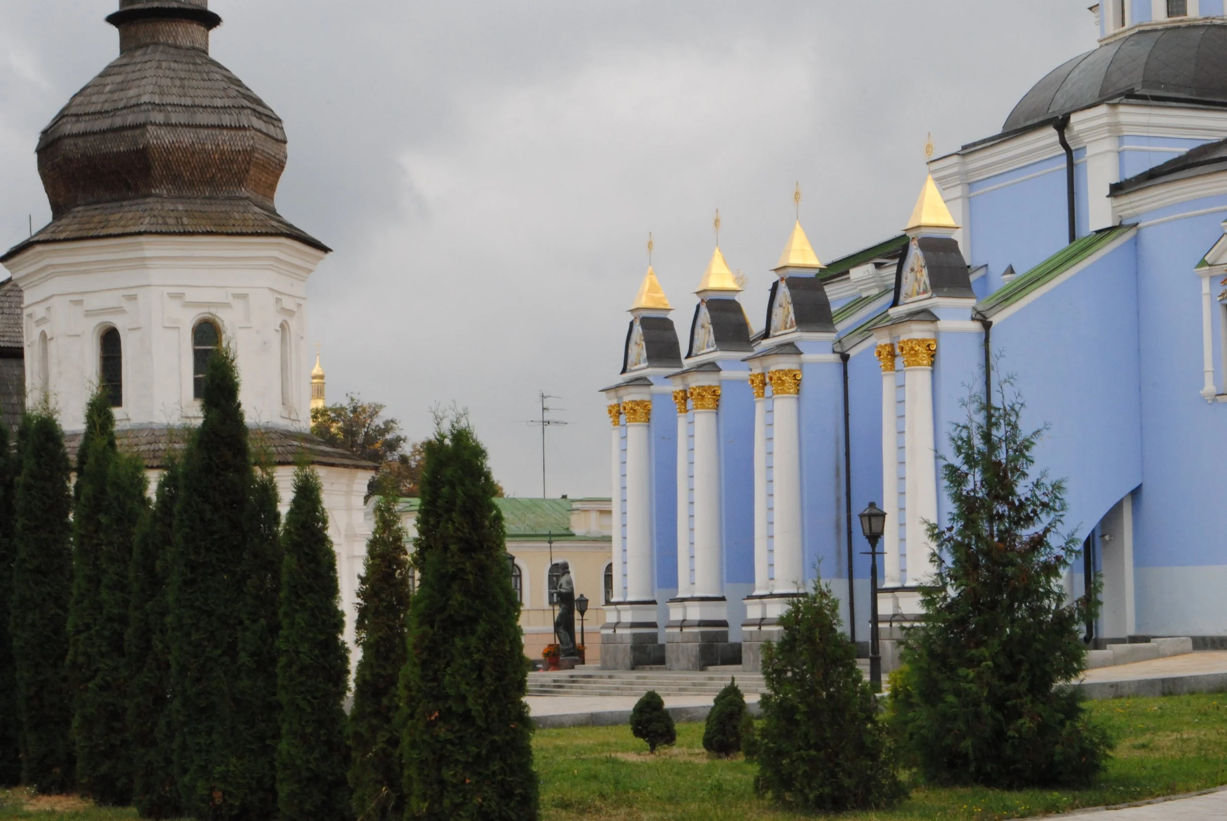 Church with white walls, gold accents, and a large domed roof, partially obscured by tall evergreen trees in a park setting under a cloudy sky. Kiev November 2011, Marina Parrish ©
