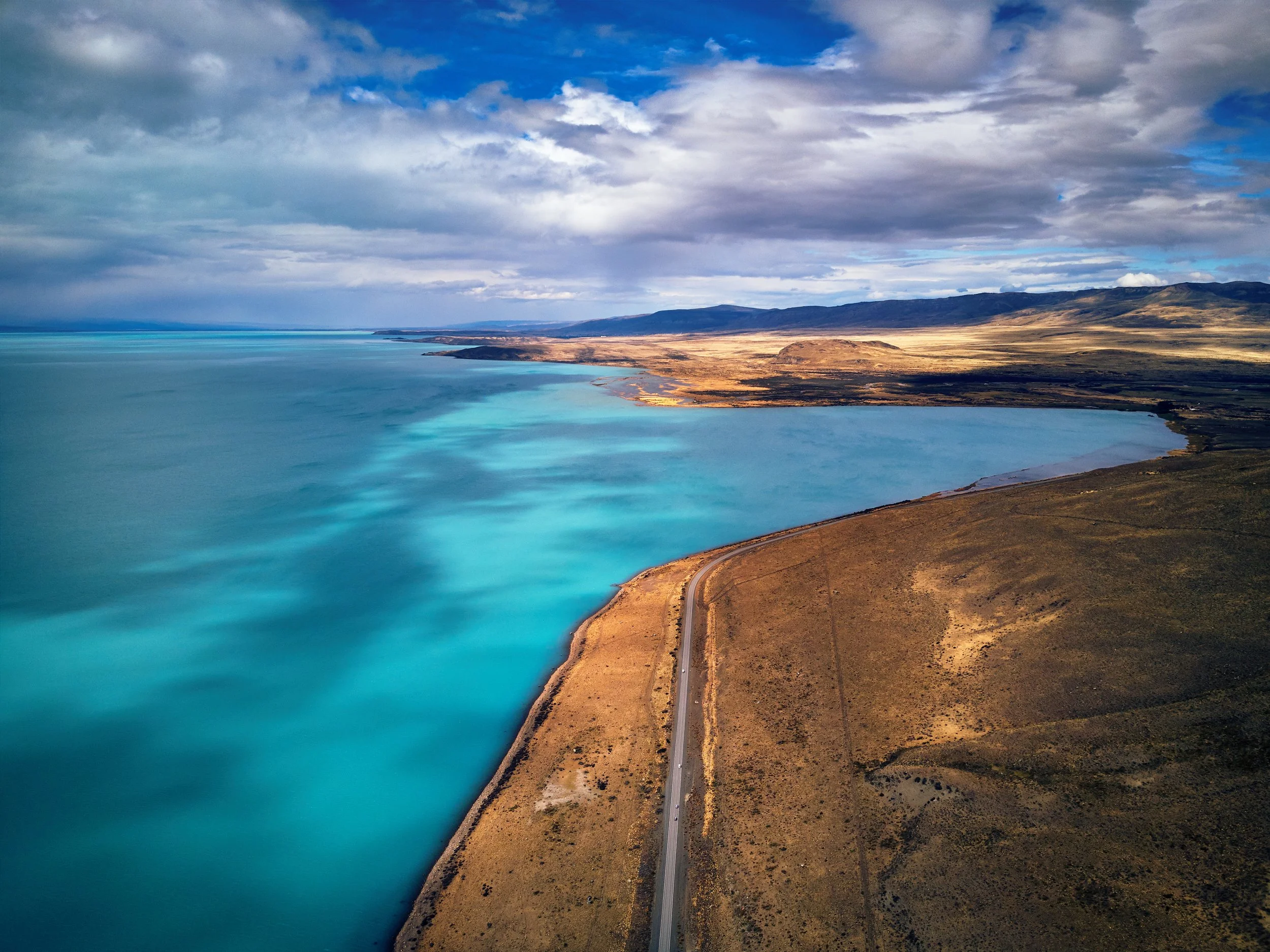 Aerial view of a large body of water with a shoreline and a road running alongside it. The landscape includes rolling hills and mountains under a cloudy sky with patches of blue.