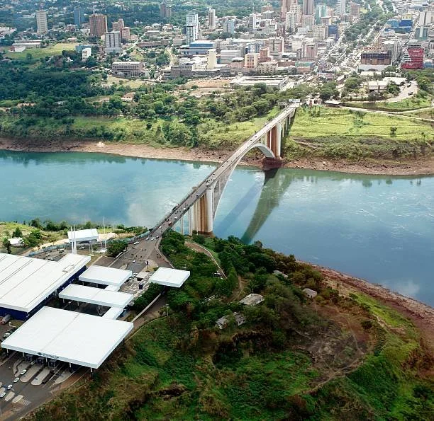 Aerial view of a city with a bridge crossing a river, surrounded by green areas and urban buildings.