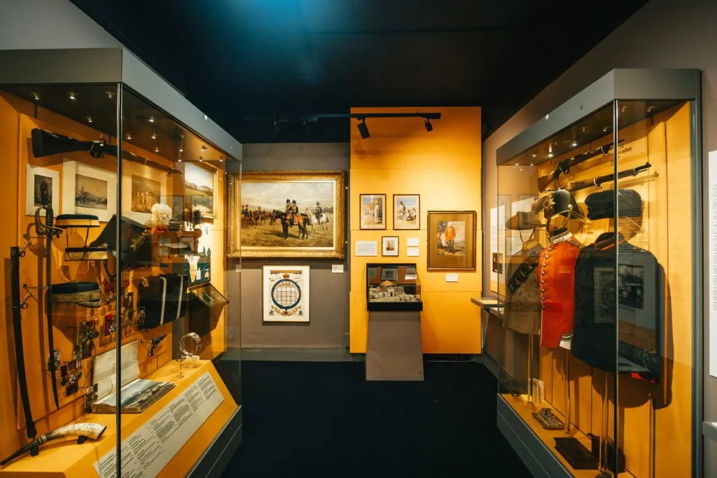An exhibit in a museum display case showcases historical military artifacts, including rifles, uniforms, and medals, with framed paintings and photographs on the walls.