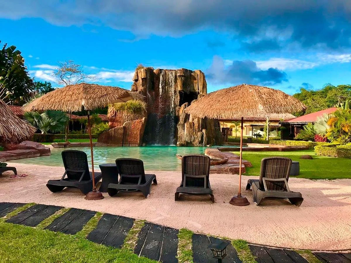 Resort pool area with lounge chairs, umbrellas, a large rock waterfall feature, and thatched roofs, surrounded by lush greenery and trees.