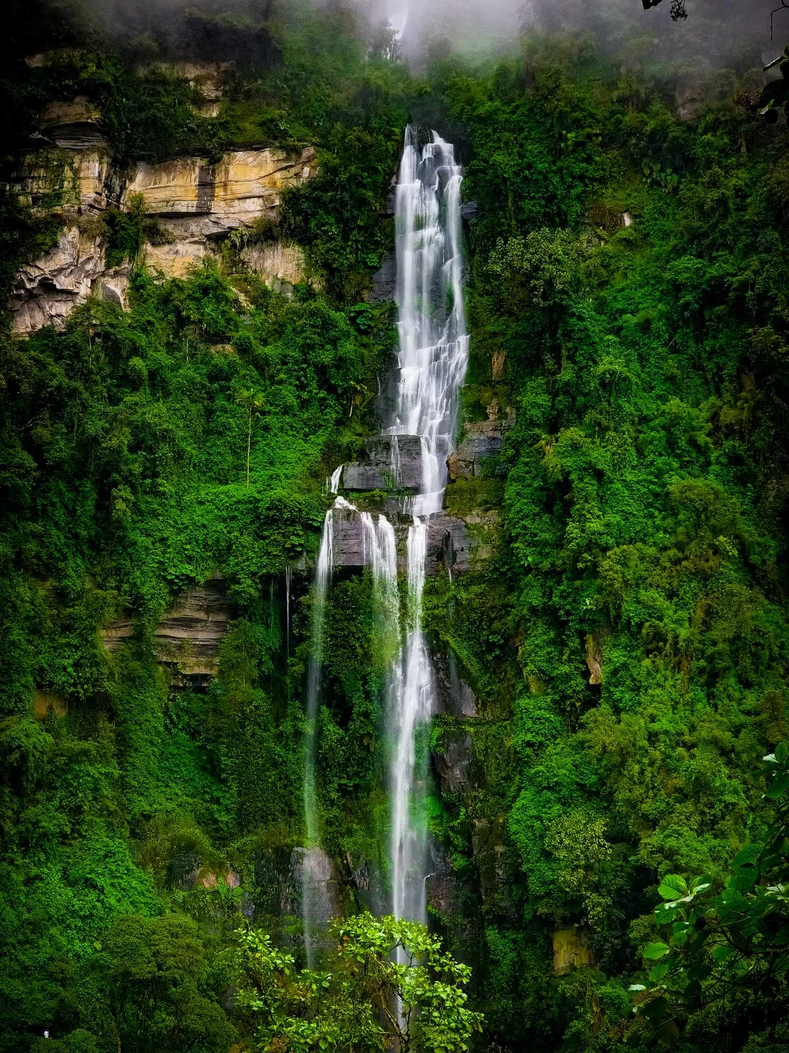 A tall, multi-tiered waterfall flowing down a lush, green, tropical forested mountainside with mist at the top.