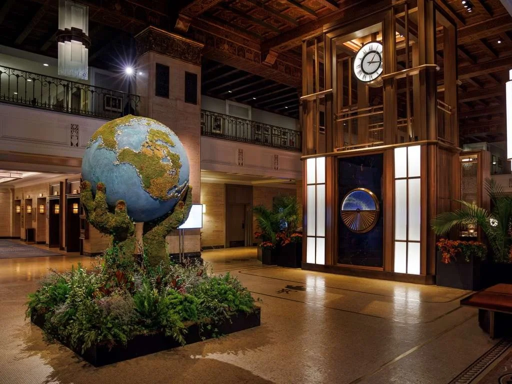 Hotel lobby with large globe sculpture surrounded by plants and a wooden clock display with a round clock face.