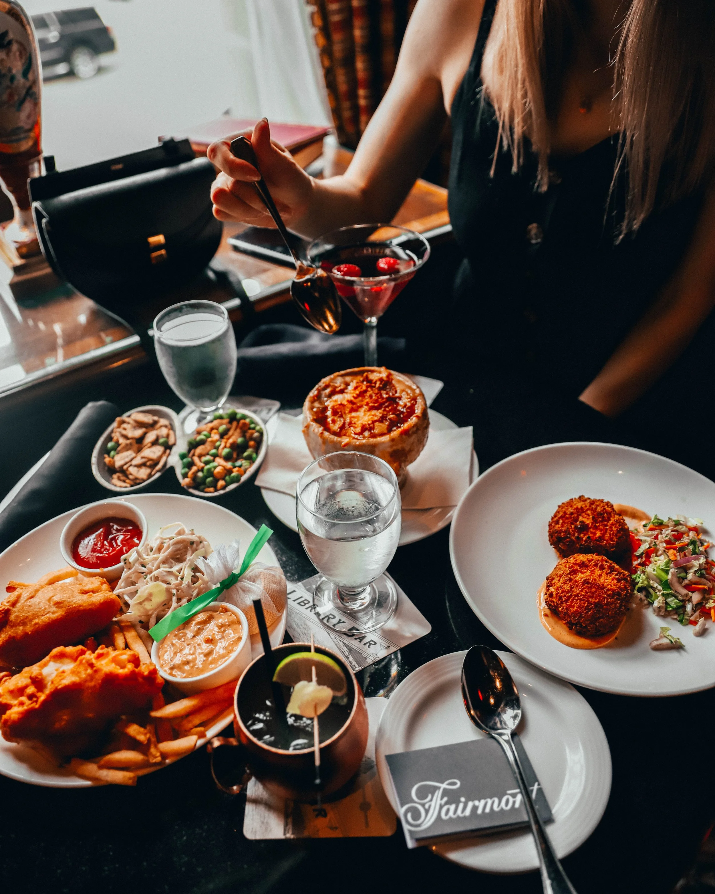 A person in a black dress is serving themselves a cherry cocktail at a dining table filled with various dishes including fried chicken, coleslaw, a baked dish, falafel with salad, desserts, and drinks.