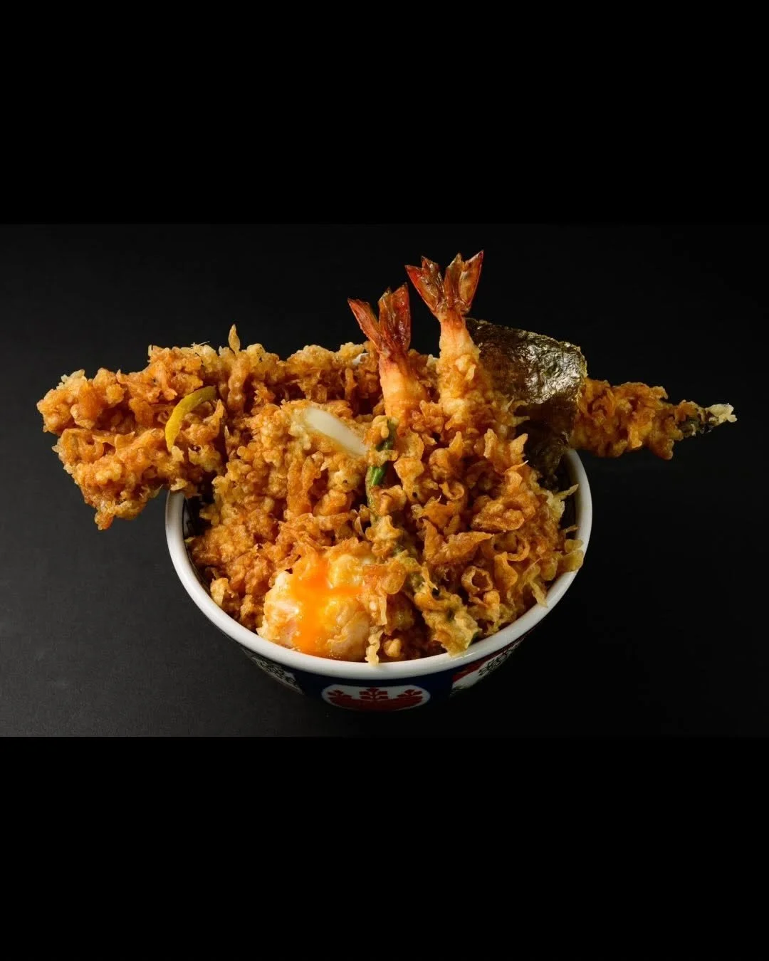 Bowl of Japanese tempura including shrimp, vegetables, and egg, served on a dark background.