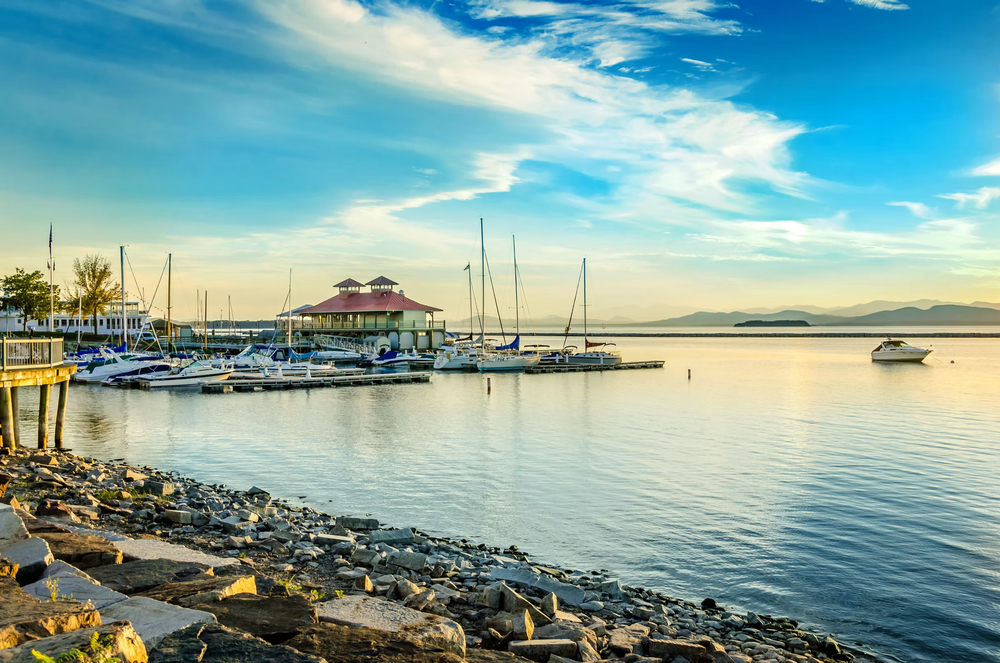 A peaceful marina at sunset with sailboats docked and a building with a red roof, rocky shoreline in the foreground, calm water, and mountains in the distance under a partly cloudy sky.