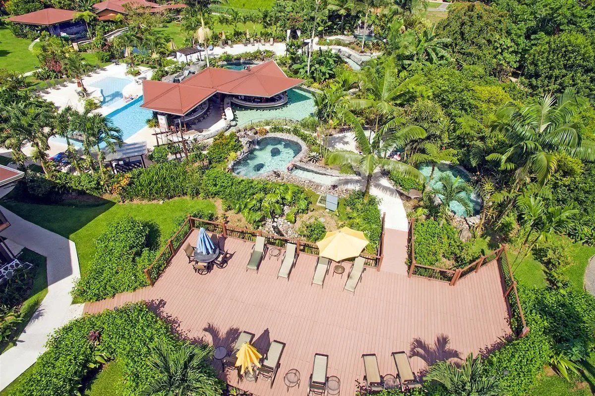 Aerial view of a tropical resort pool area with multiple swimming pools, lush green palm trees, and a large wooden deck with sun loungers and umbrellas.