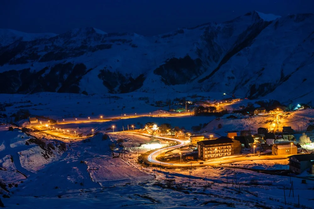 Snow-covered mountainous landscape at night with a small town illuminated by streetlights and buildings