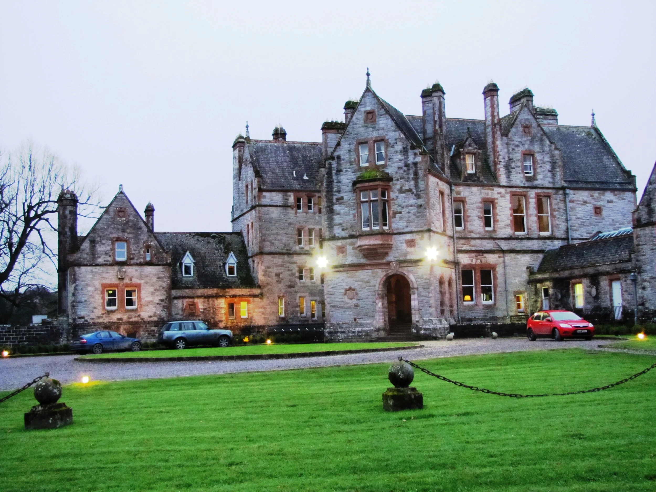 A large historic stone castle with multiple turrets and chimneys, illuminated by exterior lights, set against a cloudy evening sky. There is a parking lot with several cars in front of the castle and a well-kept grassy area with decorative chains and