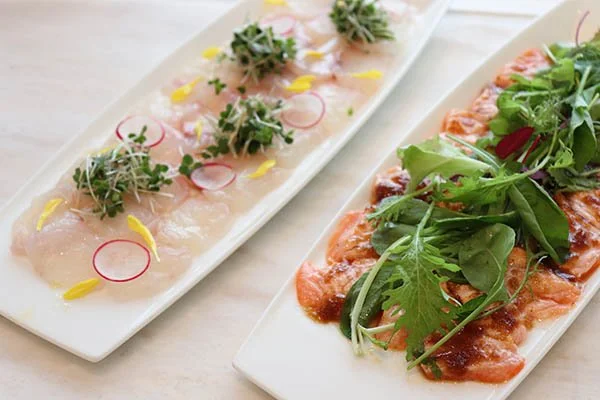 Two long white plates with thin slices of raw fish topped with herbs, radish slices, and yellow flower petals, and another with cooked meat topped with arugula and tomato sauce.