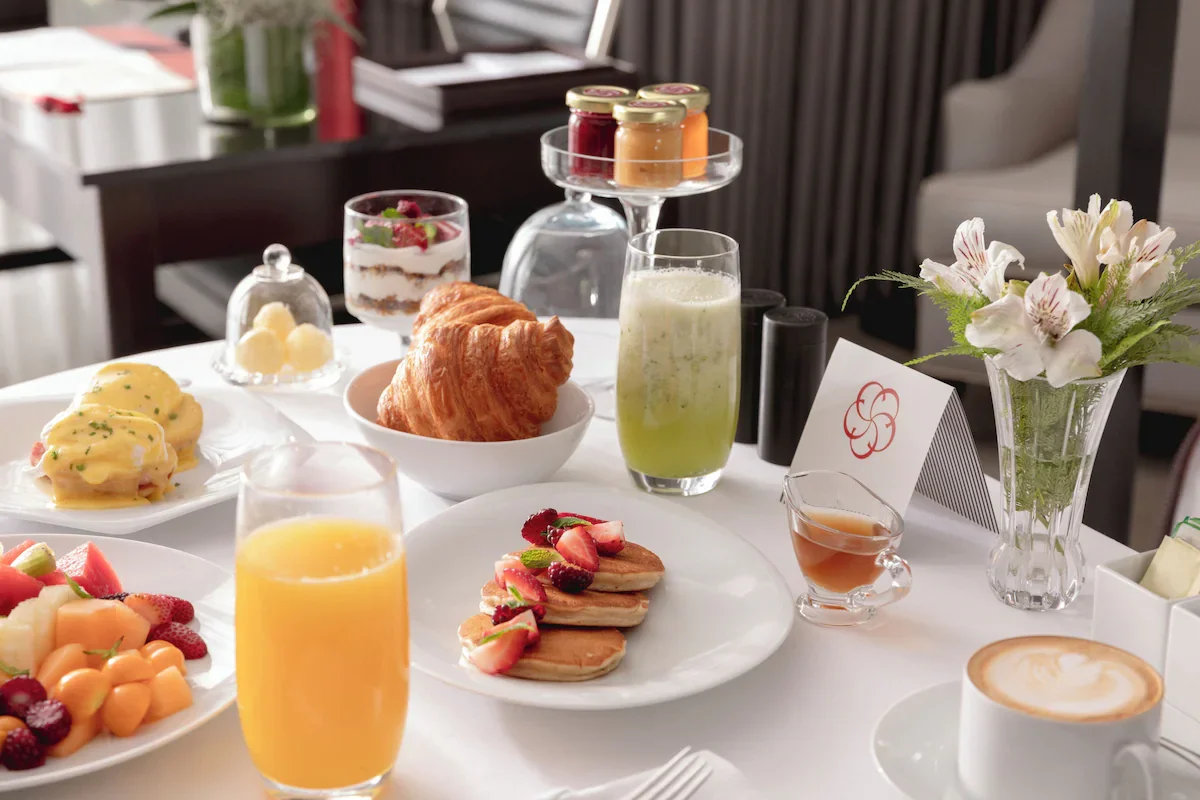 A breakfast spread with pastries, fruit, beverages, and a flower vase on a white table in a restaurant setting.