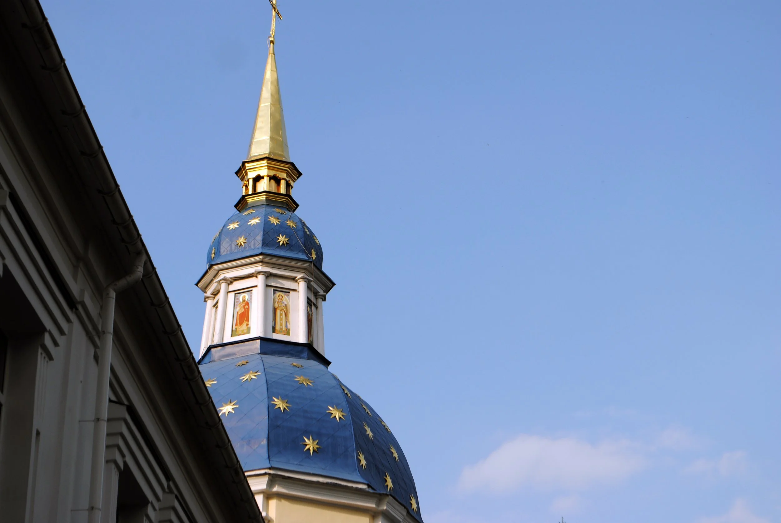 A church steeple with a blue dome decorated with gold stars, topped with a golden spire and cross, against a blue sky.