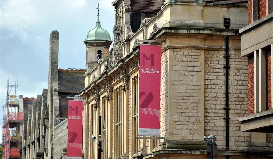 Exterior view of the Museum of Gloucester building with historic architecture and two pink banners displaying the museum's logo and name, under a cloudy sky.
