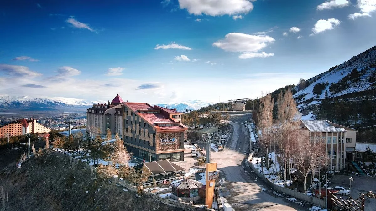 A mountain hotel with a red roof on a snowy hillside, city and mountains in the background, partly cloudy sky.
