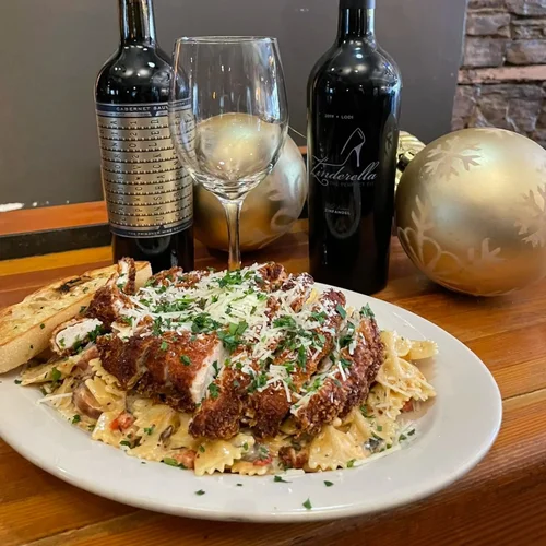 Plate of pasta with fried chicken, grated cheese, and chopped herbs, with a slice of garlic bread on the side. In the background, there are two bottles of red wine, an empty wine glass, and holiday decorations including two gold ornaments.
