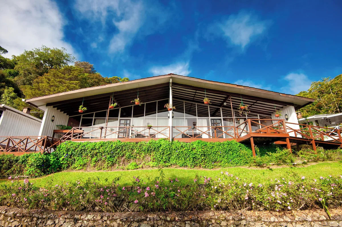 A modern house with a large glass front and wooden balcony railings, surrounded by greenery and colorful flowers, under a partly cloudy blue sky.