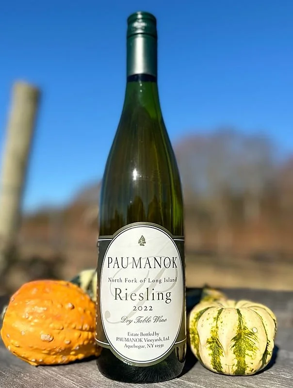 A bottle of 2022 Riesling wine from PAUMANOK vineyards, set outdoors with two decorative gourds, one orange and one green, under a clear blue sky.