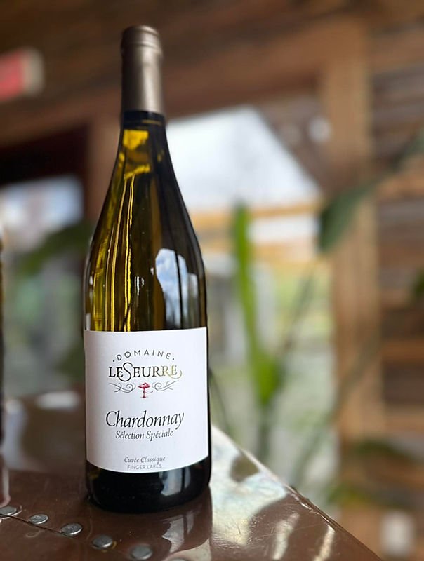 A bottle of Domaine Le Seurre Chardonnay wine on a wooden surface with a rustic wood interior and other greenery in the background.
