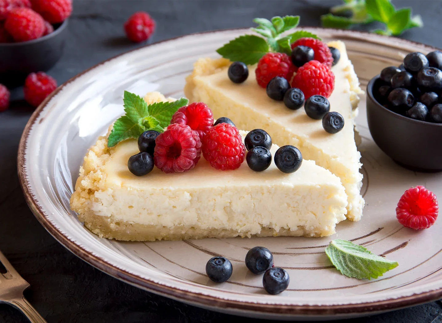 A slice of cheesecake topped with fresh raspberries and blueberries, garnished with green mint leaves on a ceramic plate.