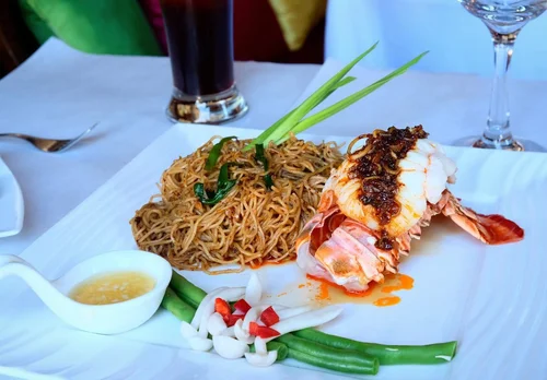 Plate of cooked noodles with green onions, a lobster tail topped with sauce, a small dish of dipping sauce, and fresh vegetables on a white tablecloth.