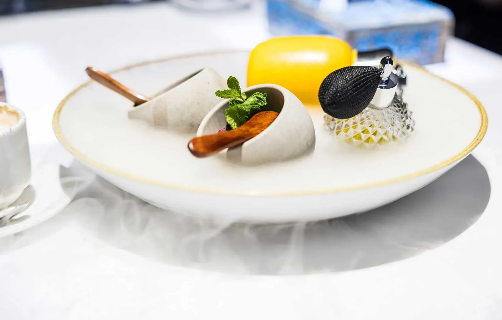 A white plate with a small cup containing green herbs, a yellow lemon, a silver and black kitchen gadget, and a glass container with a textured lid, all on a white surface.