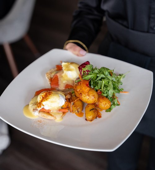 A white plate with eggs benedict with smoked salmon, roasted potatoes, and a side salad with greens and shredded carrots.