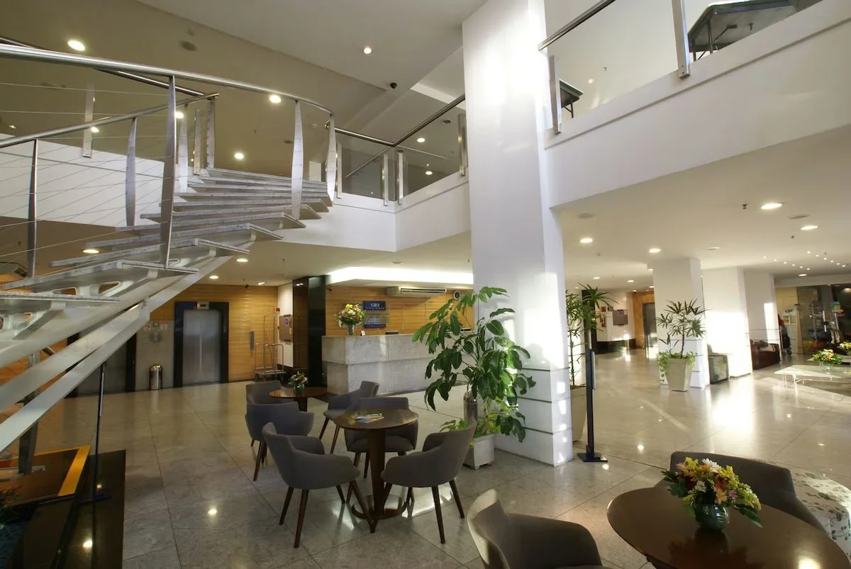 Interior of a modern hotel lobby with seating areas, potted plants, and a staircase with glass railings.