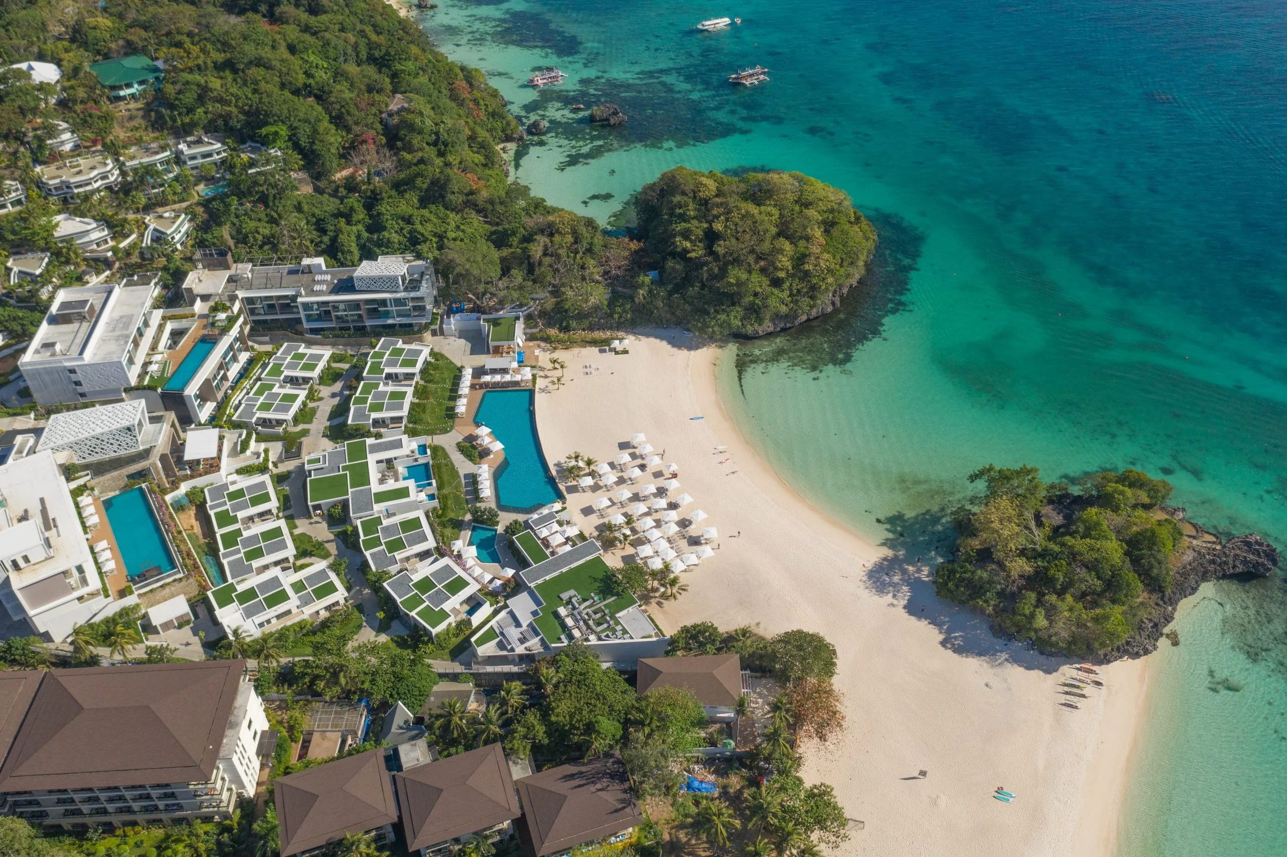 Aerial view of a tropical resort with multiple modern buildings, some with rooftop pools, surrounded by lush greenery and adjacent to a sandy beach. The clear turquoise ocean with boats is visible at the top of the image.