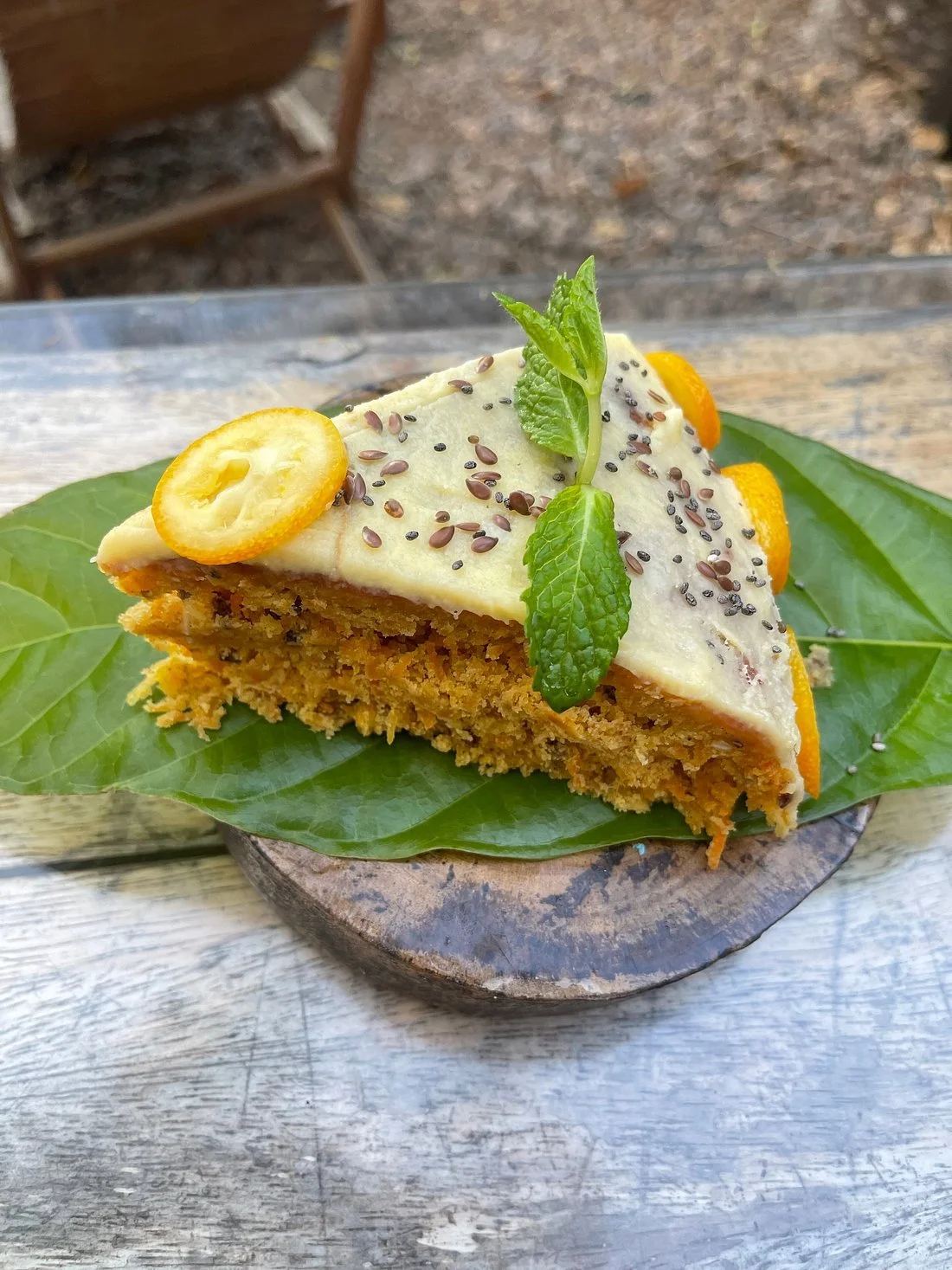 Slice of carrot cake with white frosting, garnished with lemon slices, chia seeds, and mint leaves on a green leaf.
