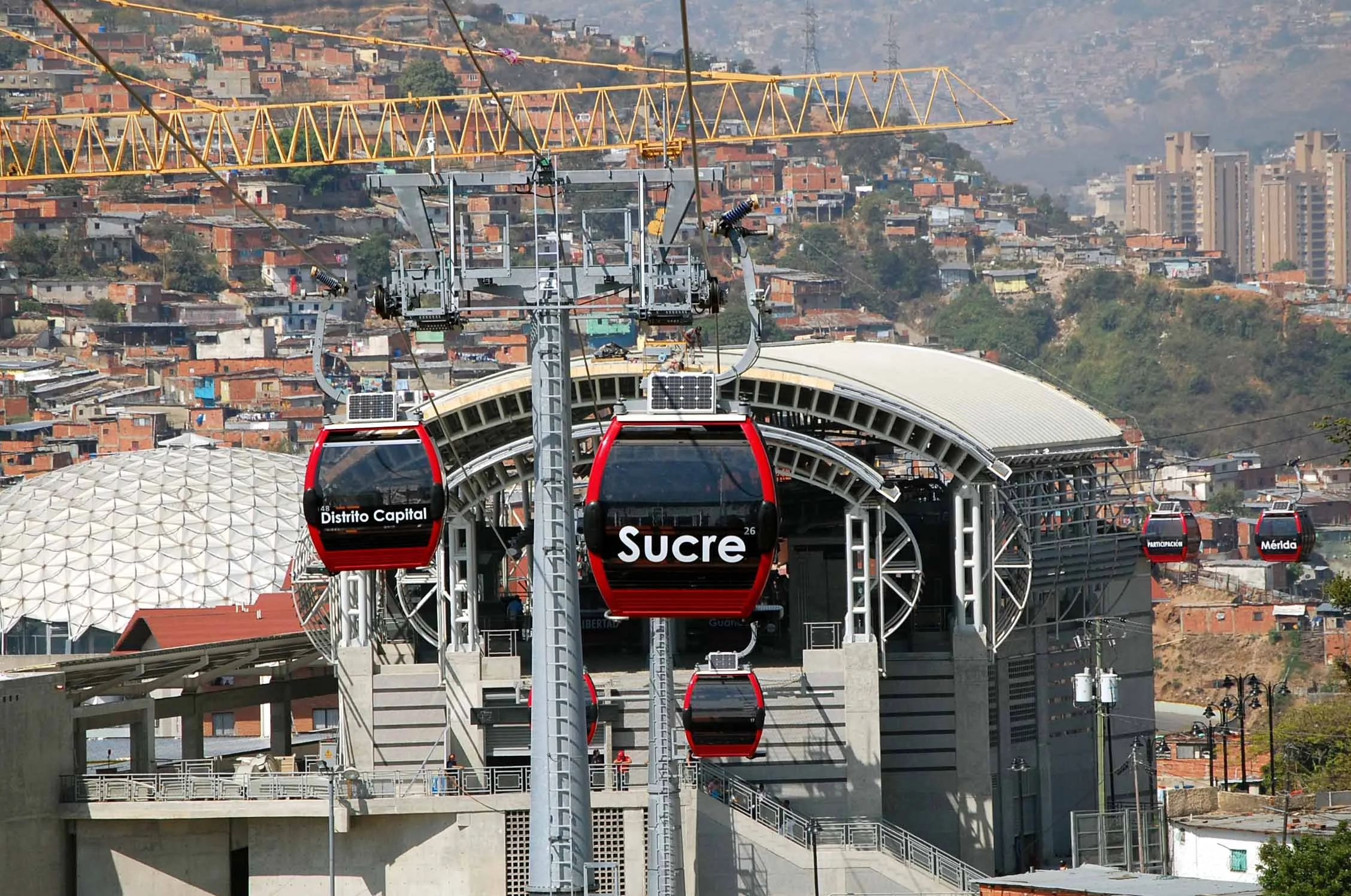 Cable car system with several red cabins labeled with station names such as 'Distrito Capital' and 'Sucre' descending from an elevated station in a densely populated urban area with hills and numerous buildings.