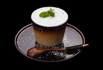 A layered dessert with a mint leaf on top, served with a spoon on a black plate, with a dusting of powdered sugar around it.