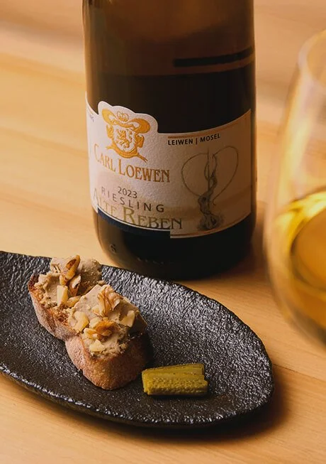 A bottle of Carl Loewen 2023 Riesling wine beside a glass of white wine, and a black slate plate with a piece of toast topped with nuts and a small piece of green wasabi, on a wooden table.