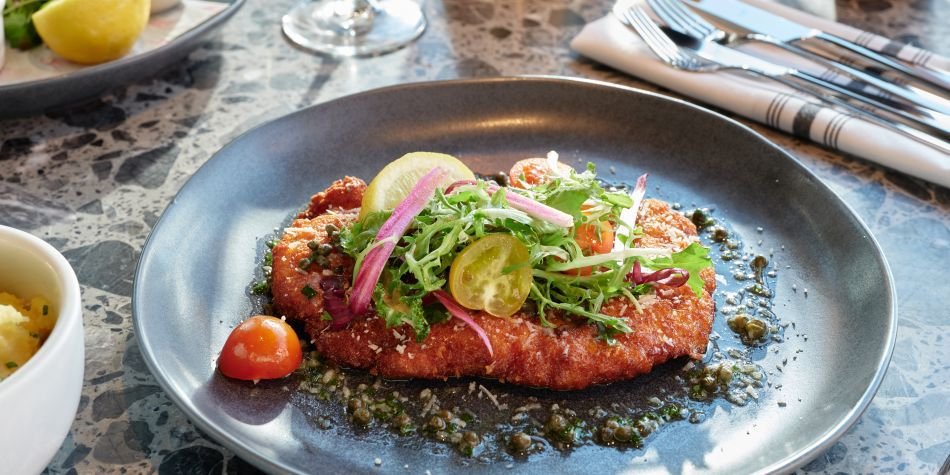 Breaded fish fillet topped with mixed greens, cherry tomatoes, and sliced cucumber on a black plate.