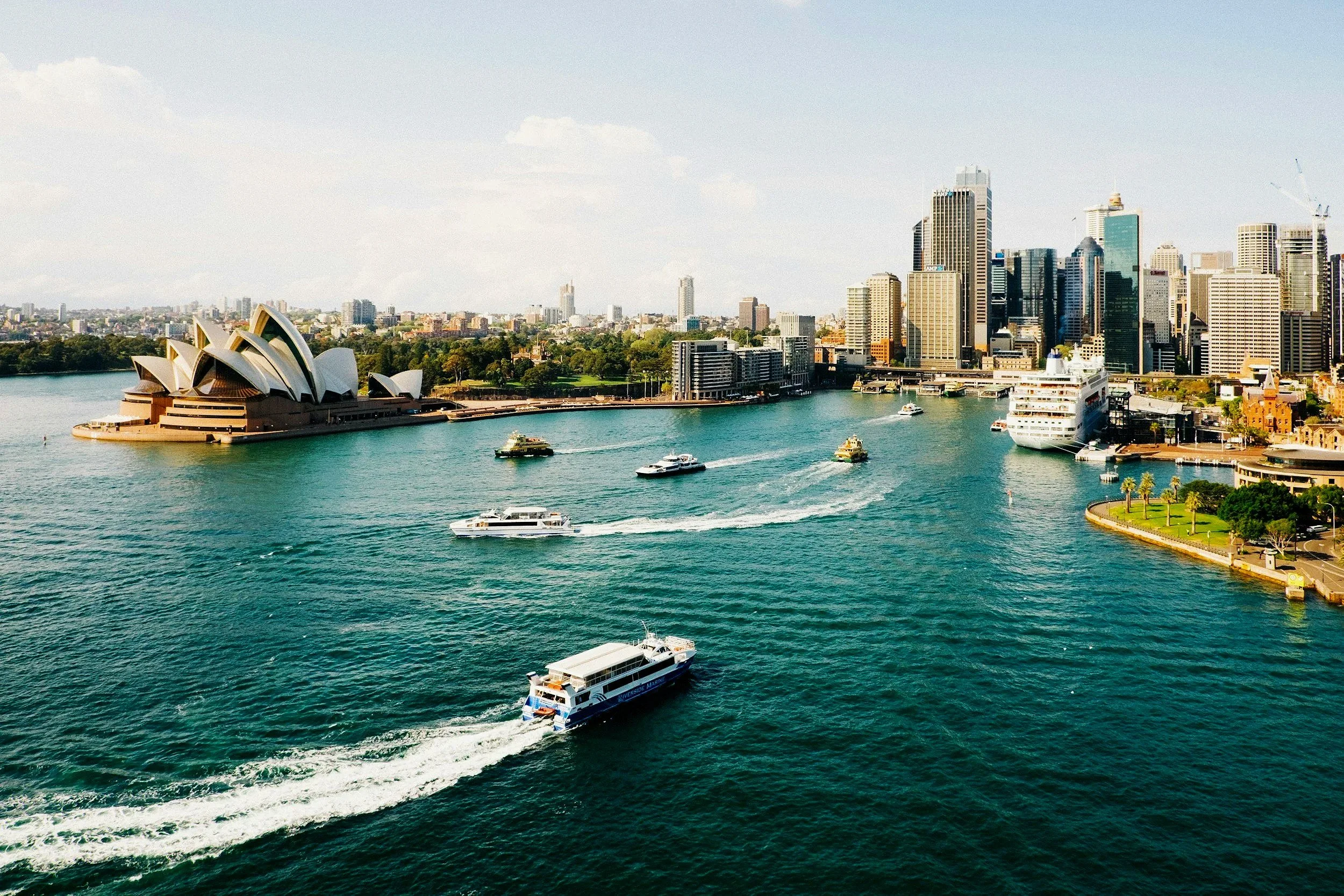 Aerial view of Sydney city skyline with the Sydney Opera House and harbor, showing boats and ferries on the water.