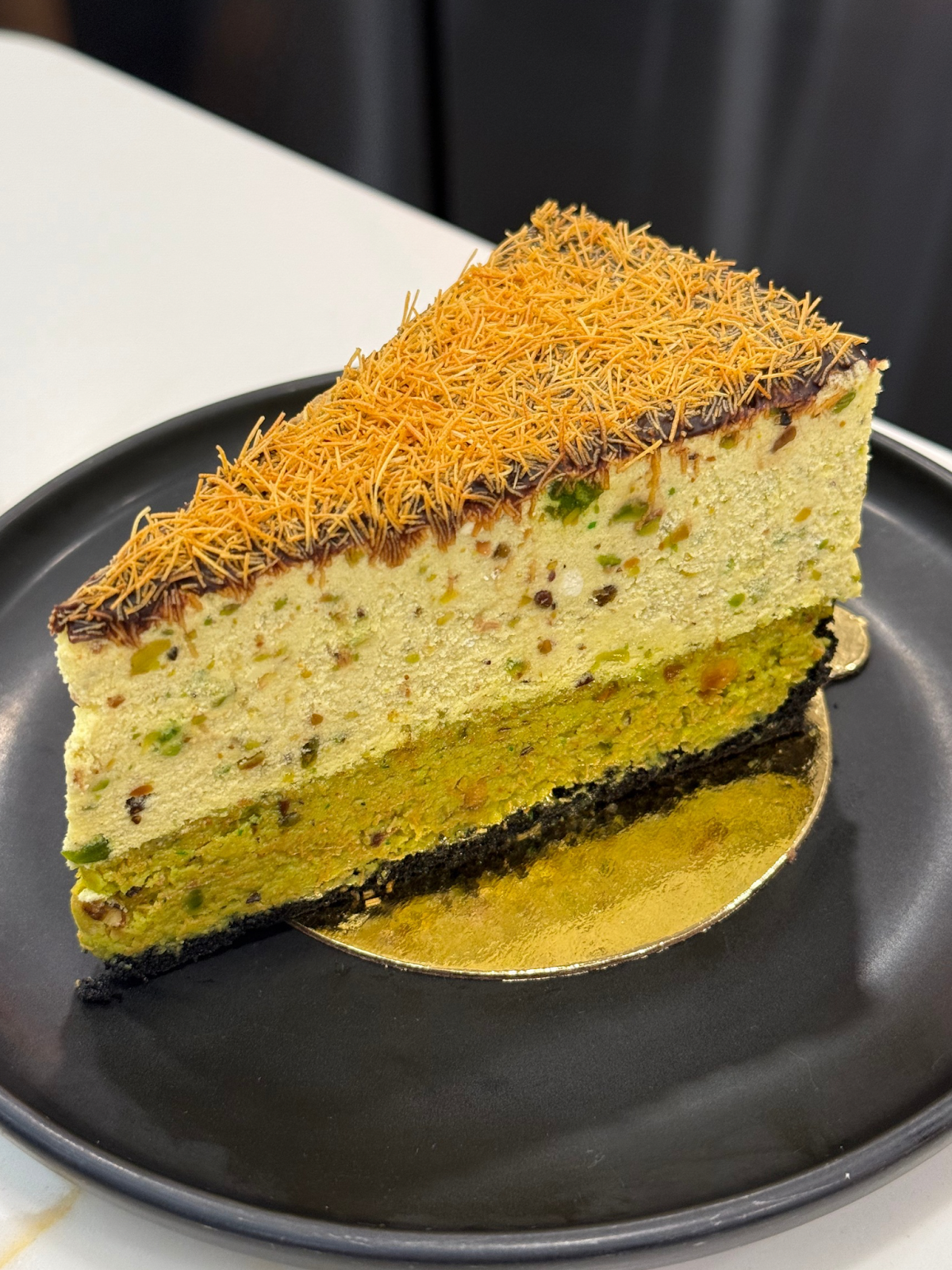 A slice of green pistachio cake with chocolate ganache and orange sprinkles on top, served on a black plate.