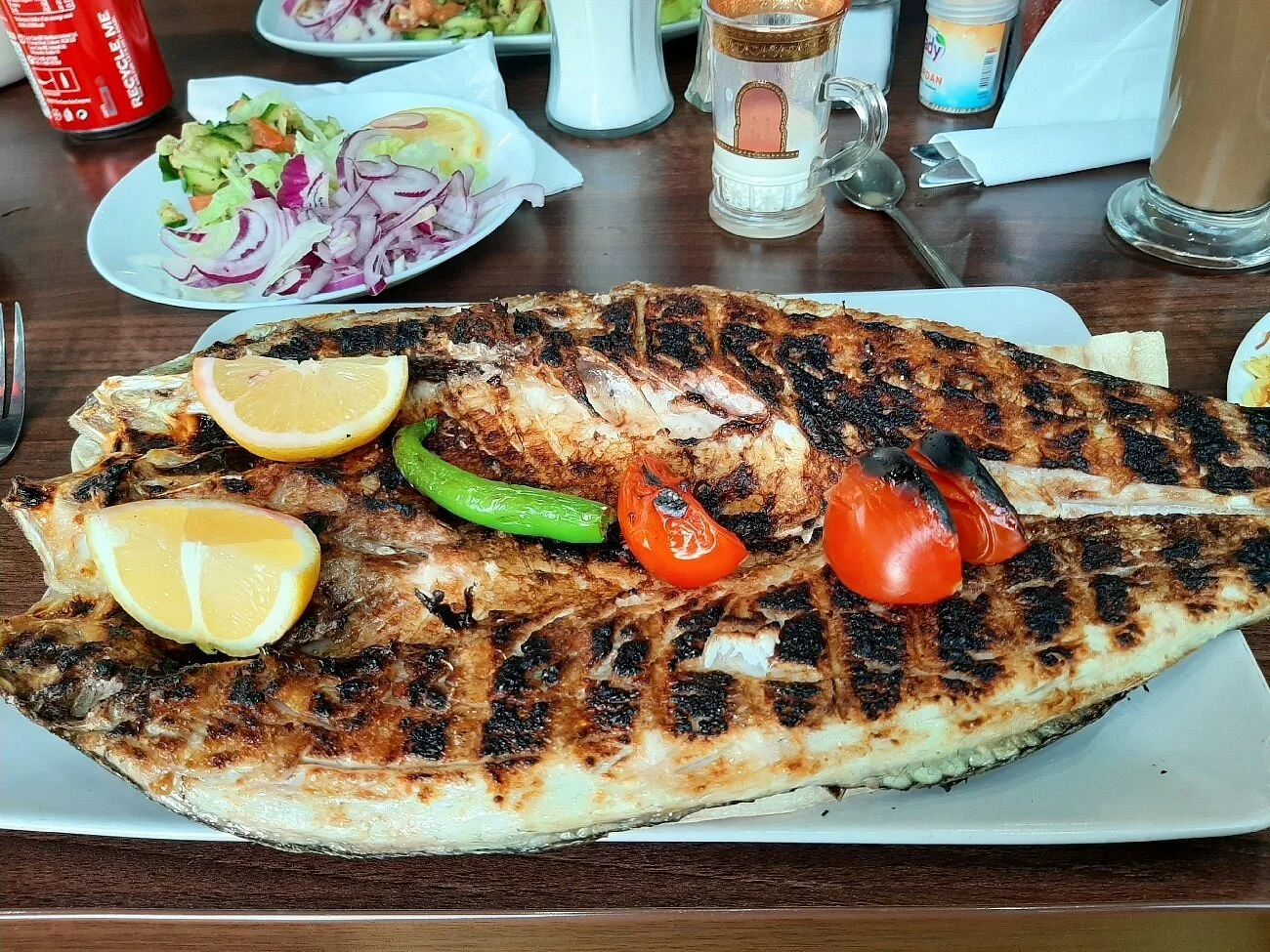 Grilled fish with lemon wedges, cherry tomatoes, and a green chili on a white rectangular plate. In the background, a salad with cucumbers and onions, and various drinks and condiments.