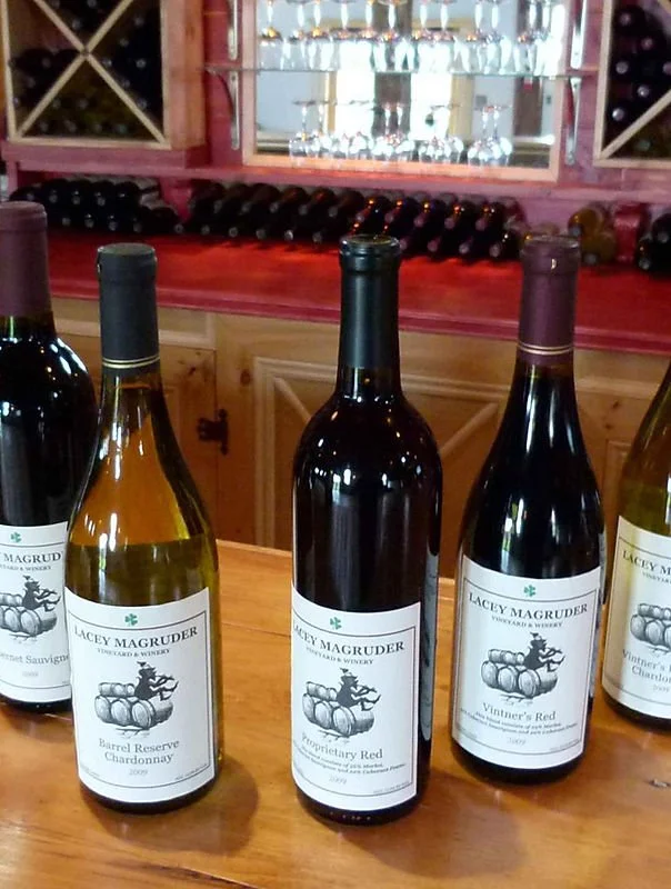 Four bottles of wine from Lacey Magruder Vineyard & Winery on a wooden counter, with a mirror and wine bottles in the background.