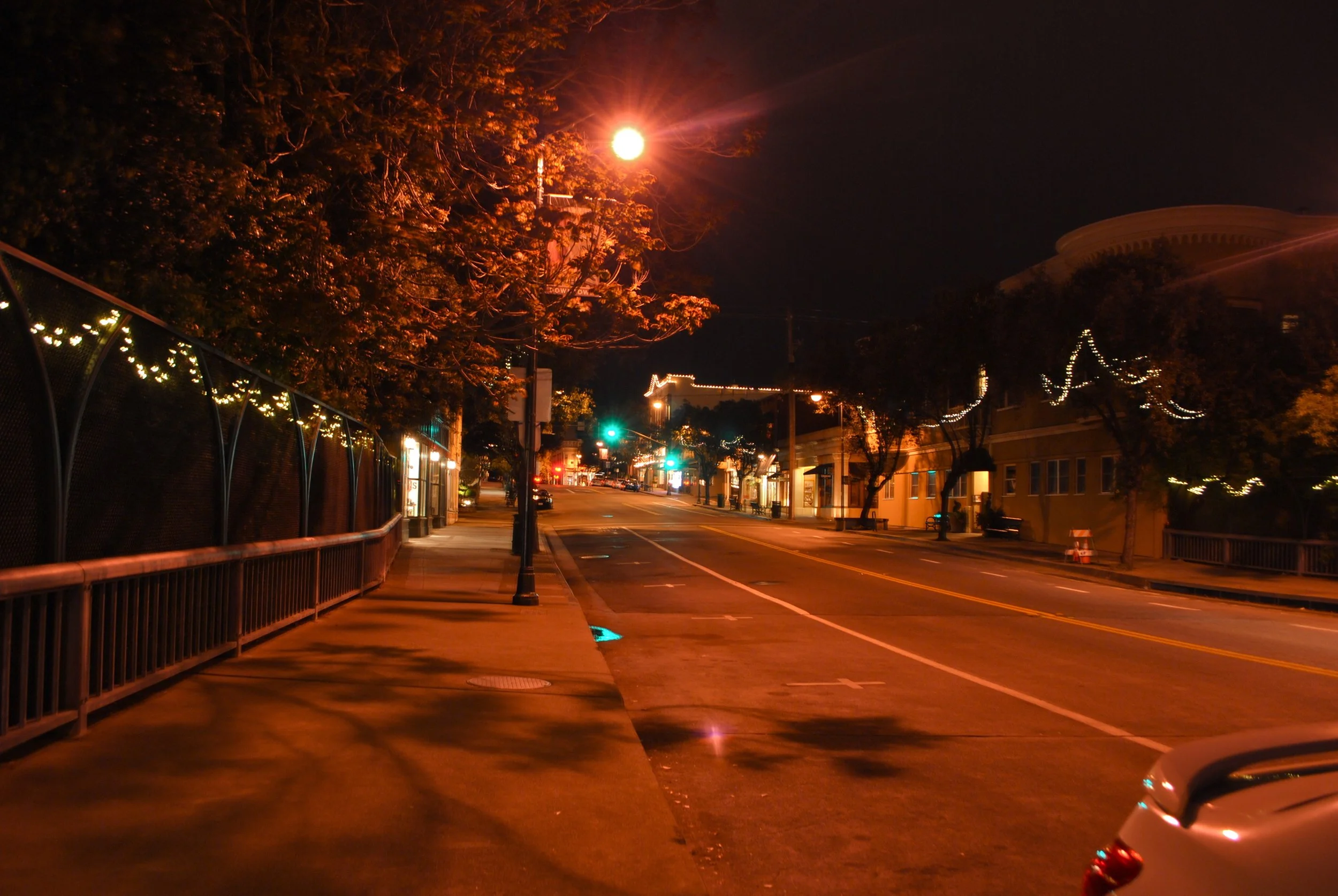 A deserted city street at night illuminated by orange streetlights, with a sidewalk, trees decorated with string lights, and buildings in the background.