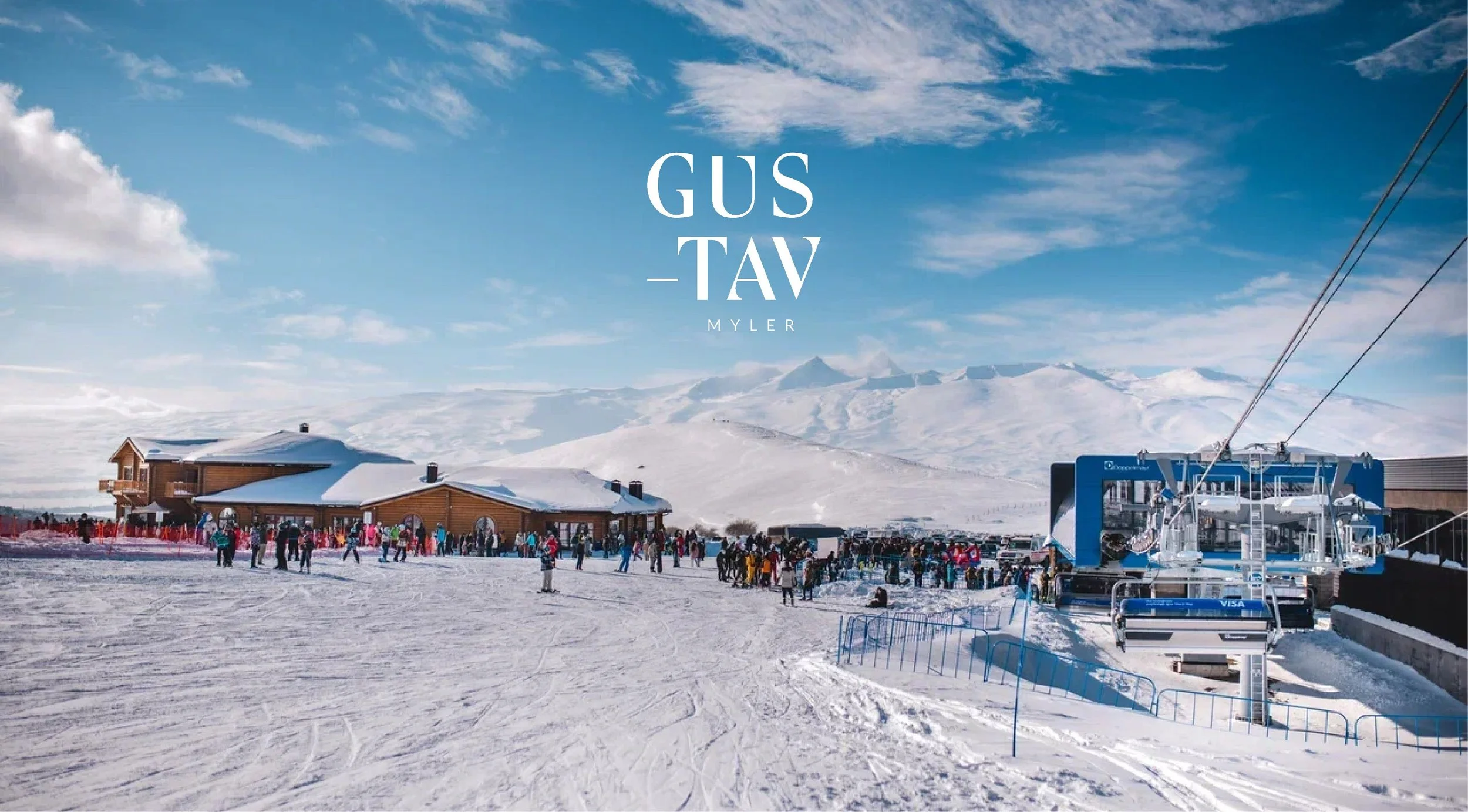 A snowy ski resort with a large wooden building, a ski lift, and many people on the snow, mountains in the background, and a partly cloudy sky.
