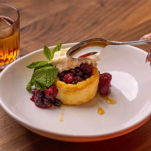A dessert featuring a cup of mixed berries inside a baked, golden crust, topped with a dollop of whipped cream, a mint sprig, and drizzled with honey, served on a white plate, with a glass of iced tea in the background.