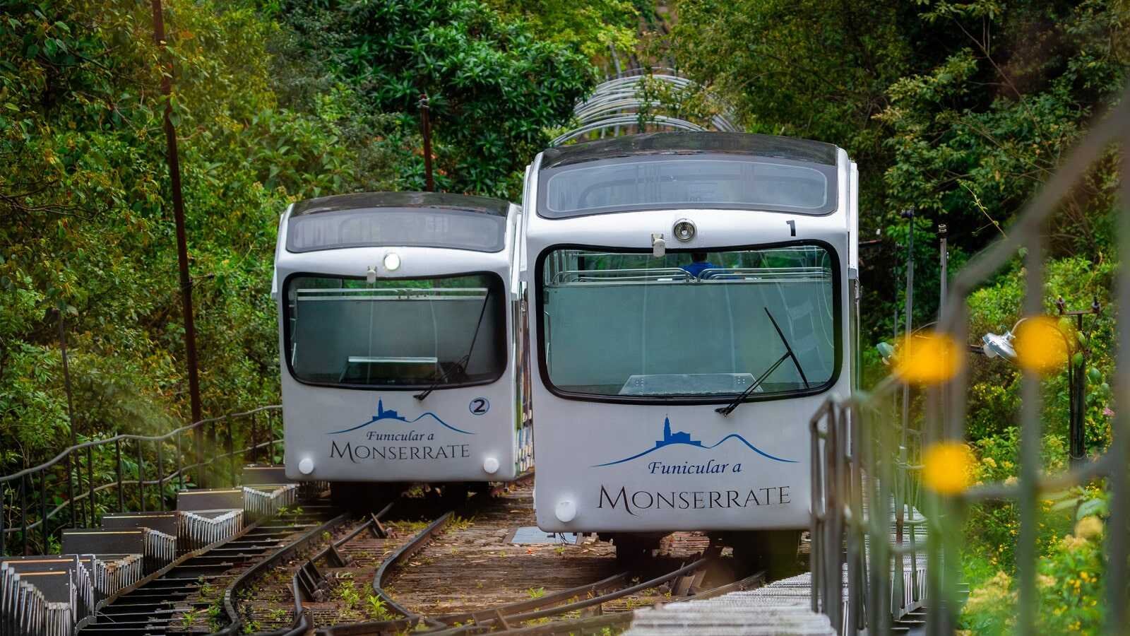 Two monorail cars on a track surrounded by green trees, with the words "Funicular a Monserrat" written on the fronts of the trains.