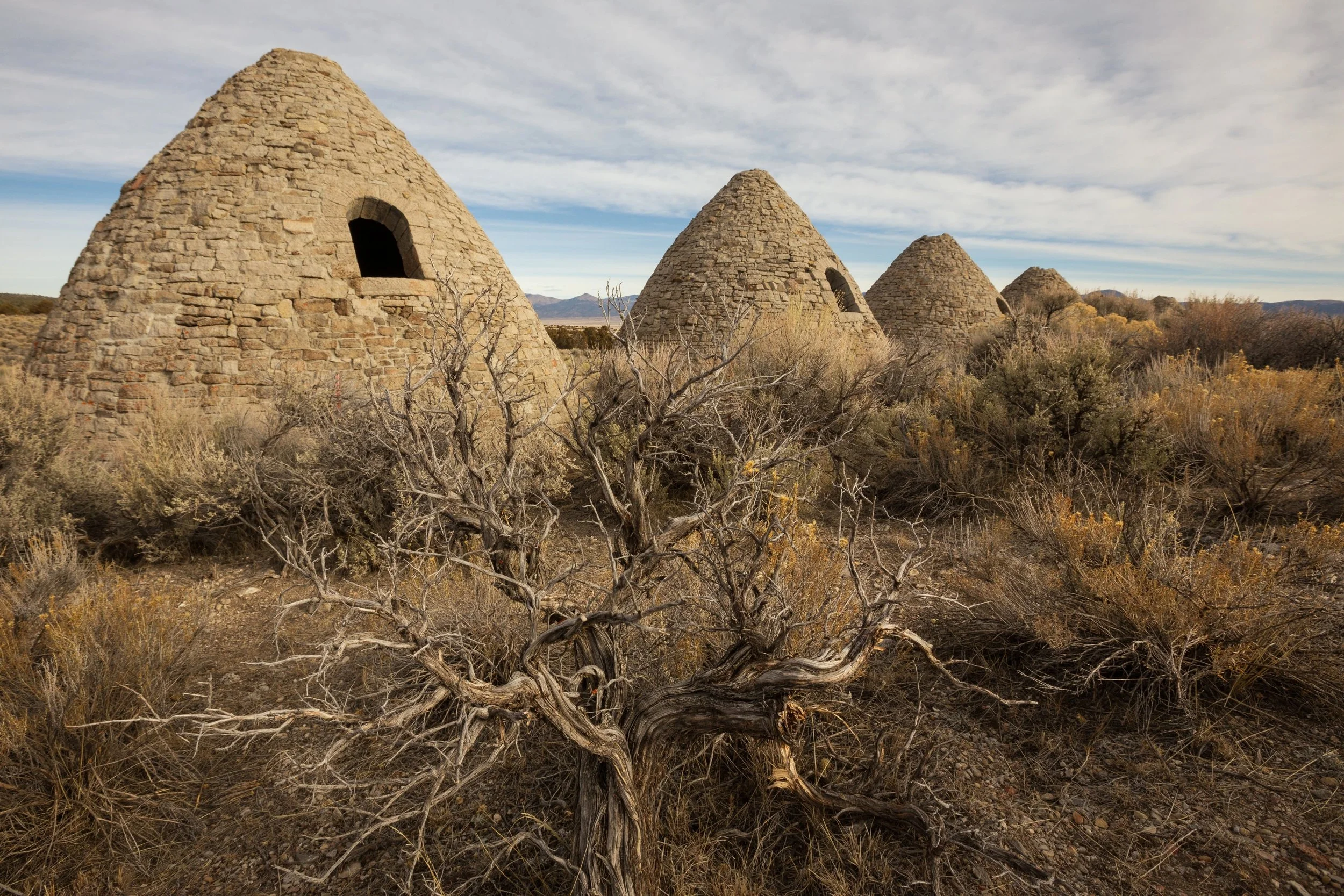 Four stone beehive-shaped structures in a desert landscape with dry bushes and mountains in the background.