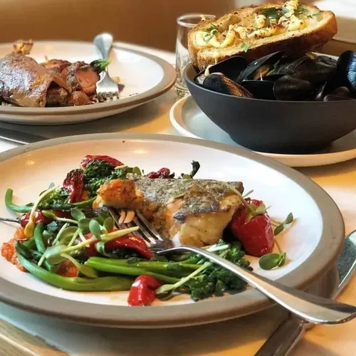 Plate with a baked fish fillet topped with vegetables and herbs, surrounded by greens, cherry tomatoes, and a fork. In the background, there are bread with melted cheese and a bowl of mussels with a bread slice resting on top.